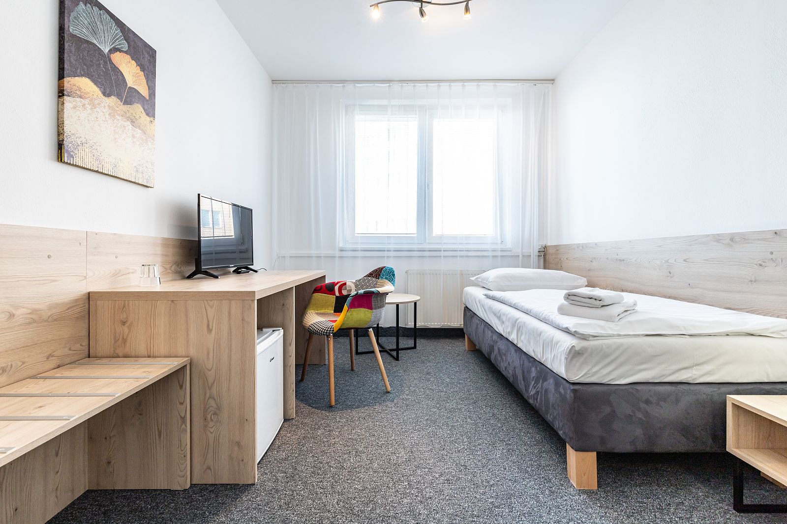 Světlem zalitý jednolůžkový pokoj s barevnou patchwork židlí u okna a mini lednicí pod stolem